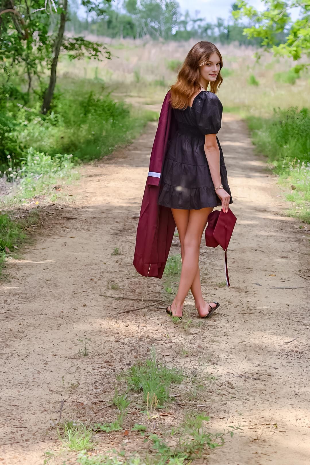 Young woman in black dress holding burgundy graduation gown walks down a rural dirt path surrounded by green trees.