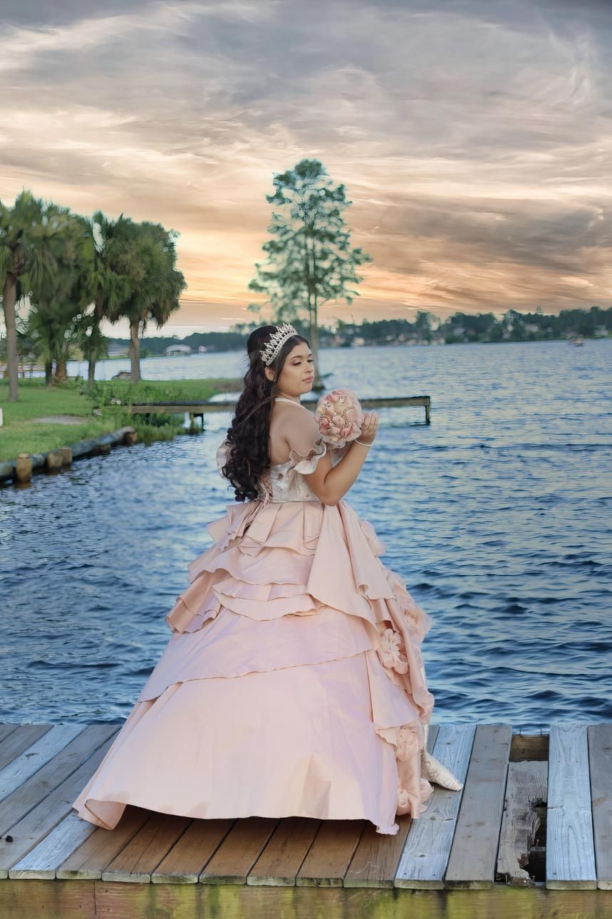 Young woman in a blush pink ball gown and tiara poses on a dock at sunset by a serene lake.