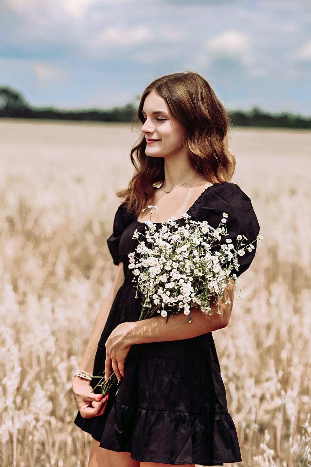 A figure in a black dress stands in a golden wheat field holding delicate white baby's breath flowers.