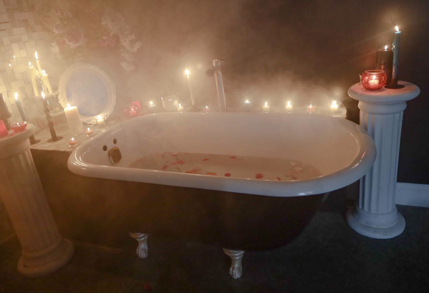 Romantic clawfoot bathtub surrounded by glowing candles and soft mist in a dimly lit, elegant bathroom.