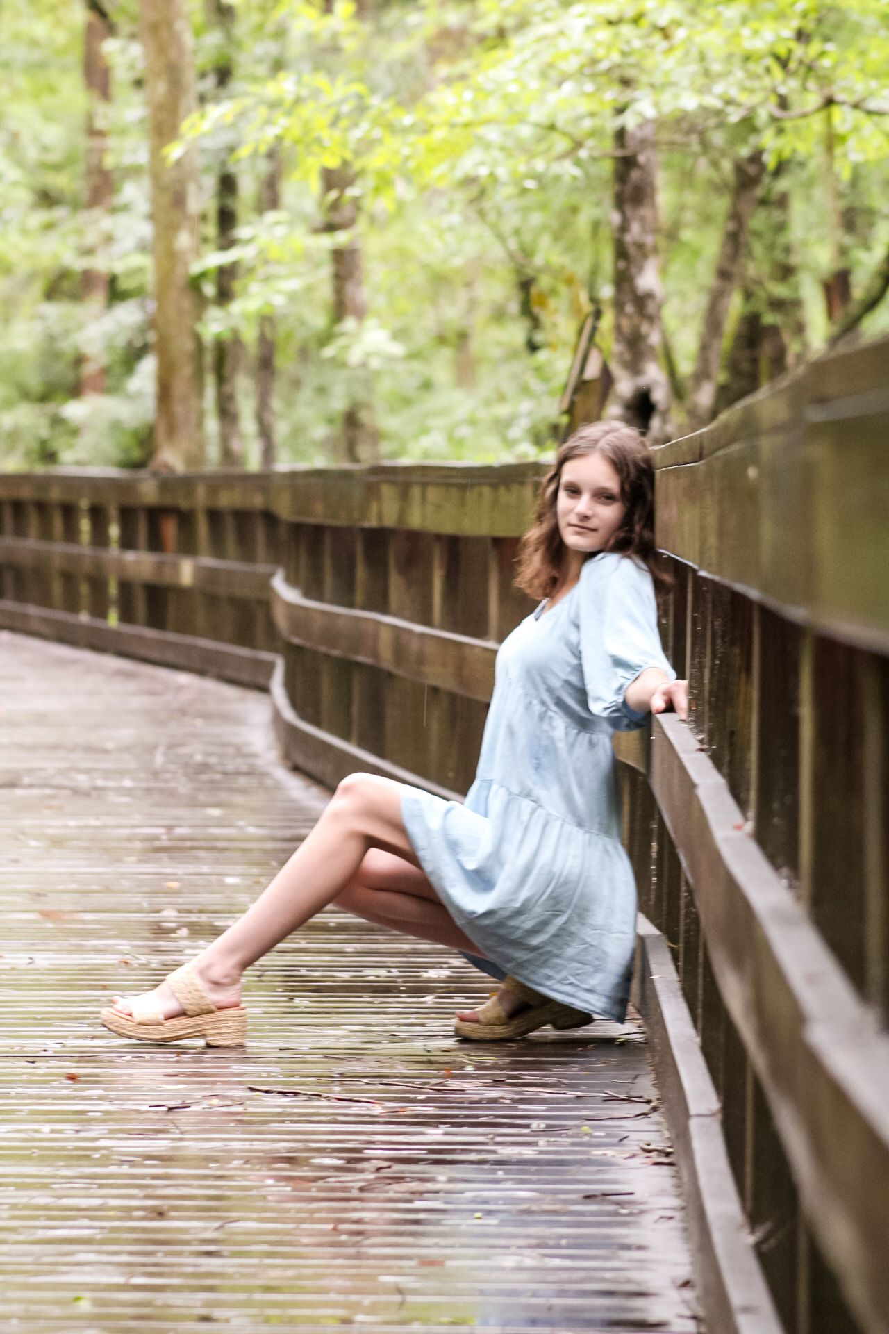 Young woman in light blue dress sitting on wooden bridge in lush green forest setting.