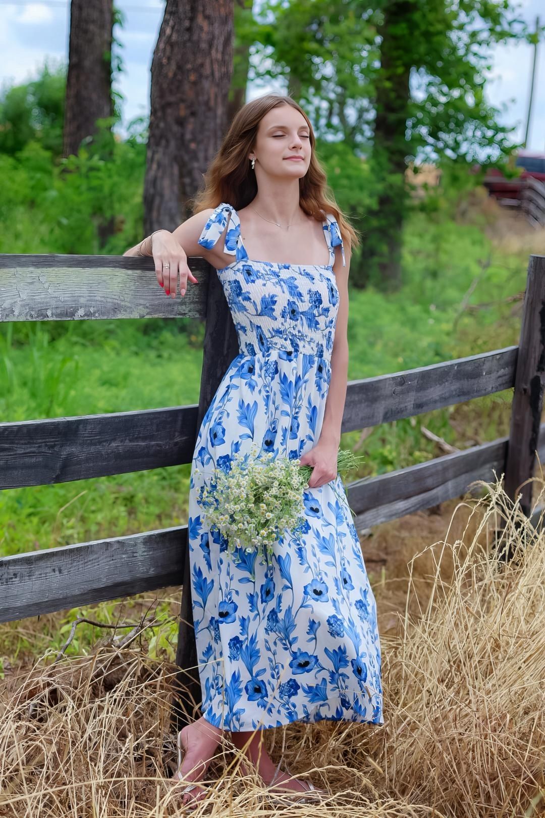 Young woman in blue floral midi dress holding wildflowers, leaning on rustic wooden fence in countryside setting.