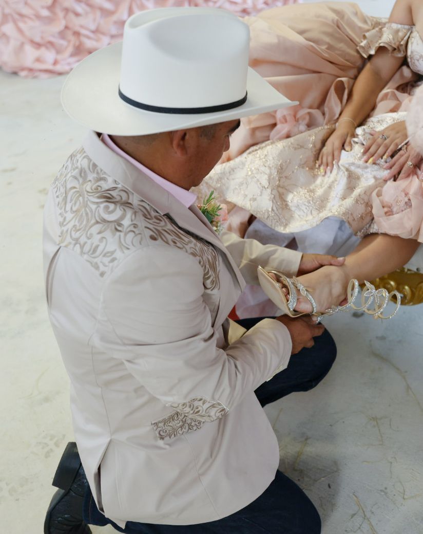 Man in white cowboy hat and embroidered jacket kneeling to place a shoe on a woman in a pink gown.