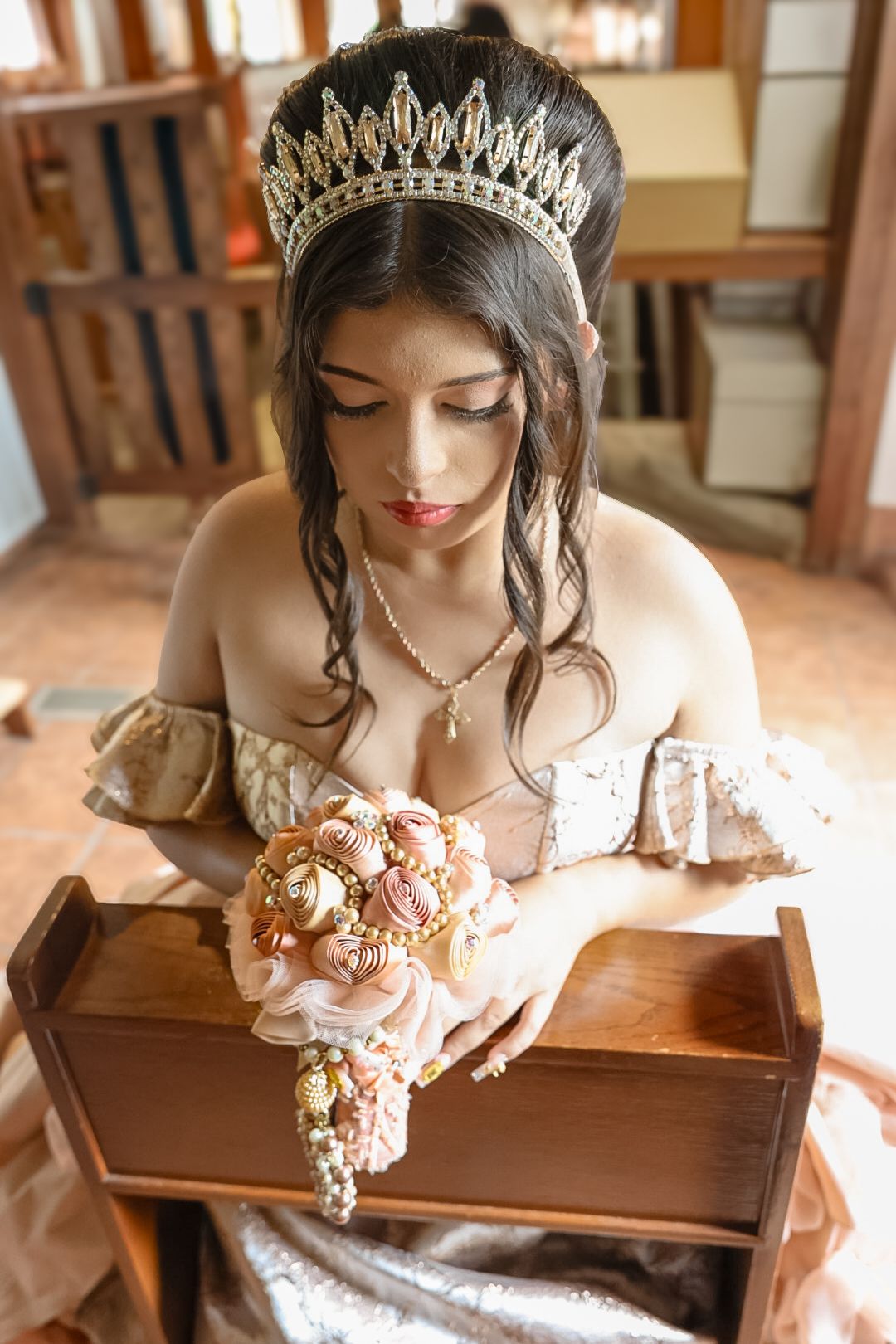 A brunette woman wearing a gold crown and off-shoulder gown holds an ornate floral bouquet while seated indoors.