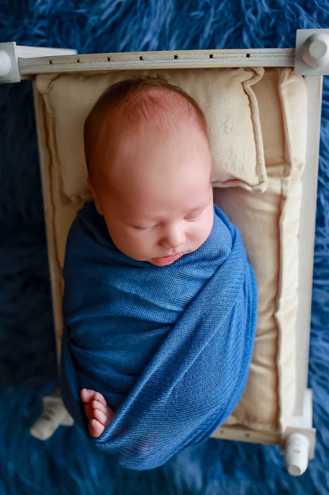 A sleeping newborn baby wrapped in a blue swaddle blanket rests in a wooden crate.