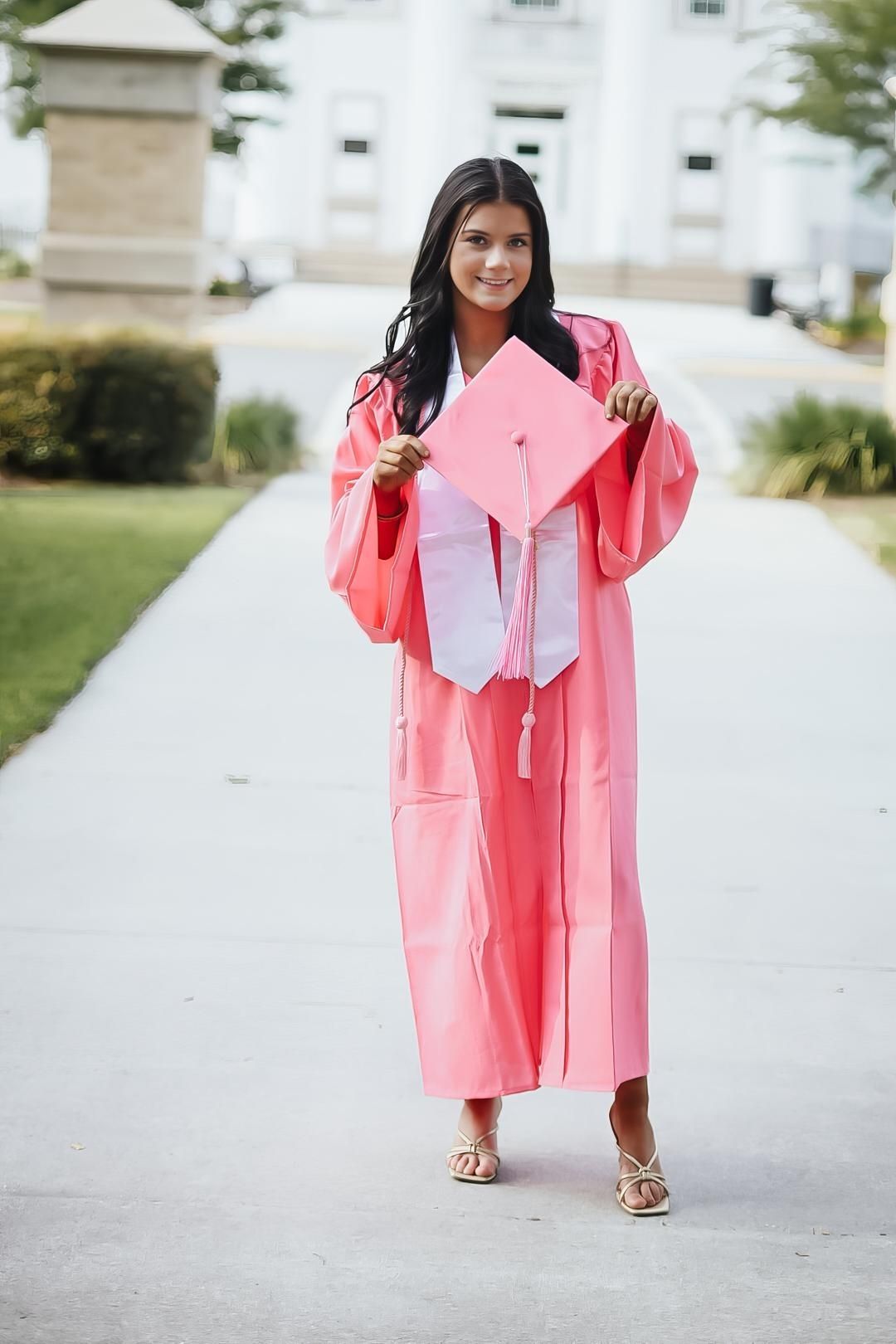 Graduate in coral pink graduation gown holds up matching cap while standing on campus walkway.