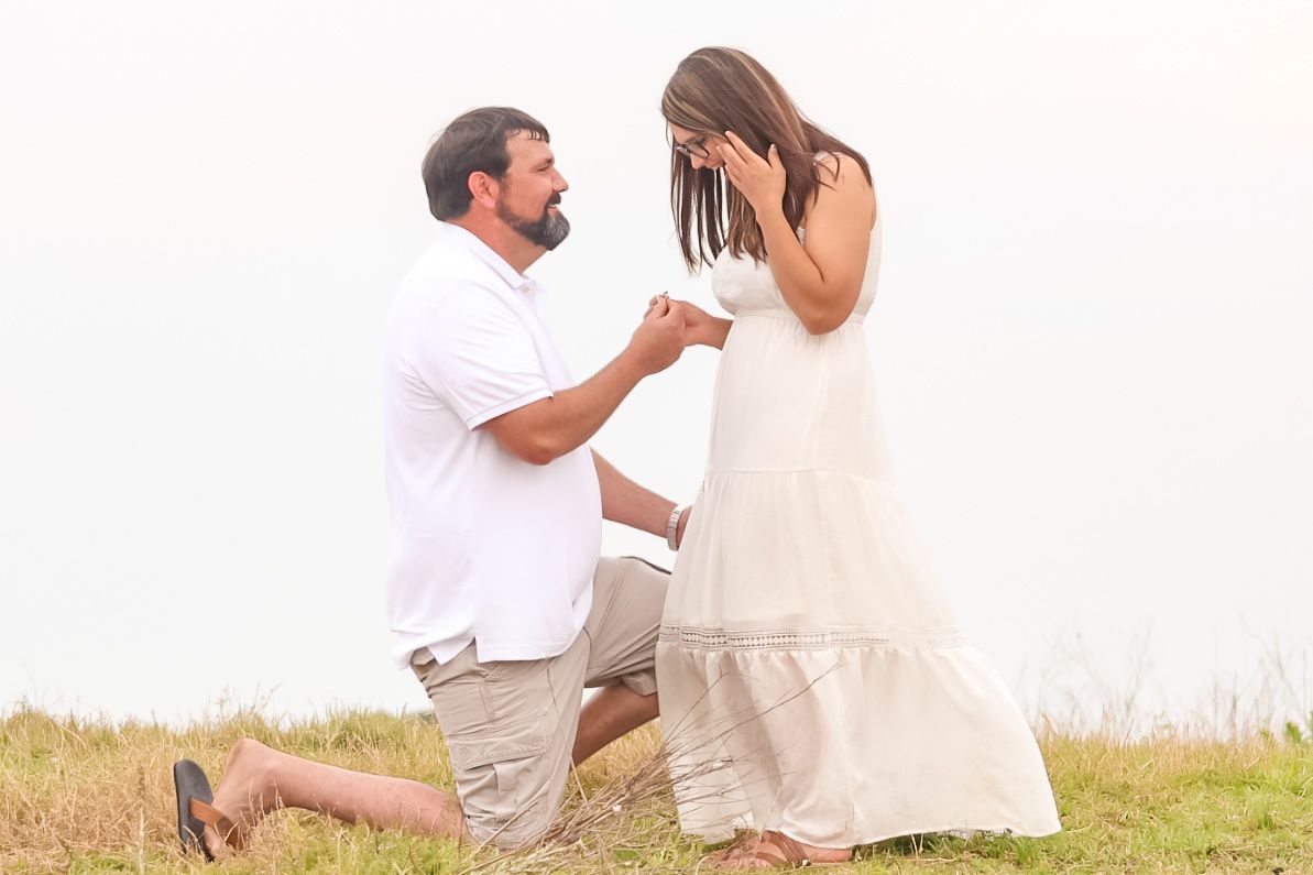 Man kneeling on grass proposing to emotional woman in white dress outdoors during golden hour engagement moment.