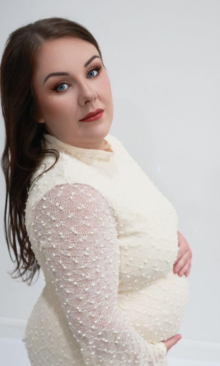 A young woman in a cream pearl-embellished dress poses elegantly against a white background with bold makeup.