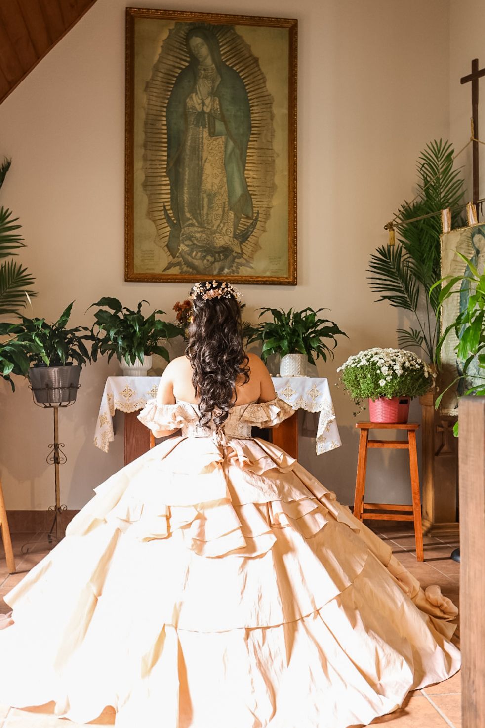 Young woman in elegant golden quinceañera ball gown kneeling before Virgin of Guadalupe altar in chapel.