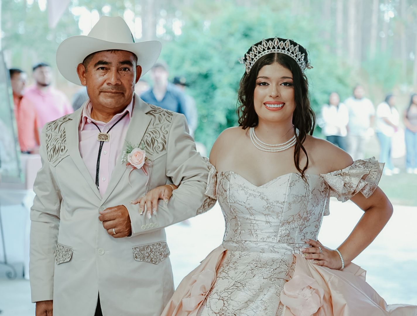 A young woman in a quinceañera gown and tiara poses with a man in a white cowboy hat at an outdoor celebration.