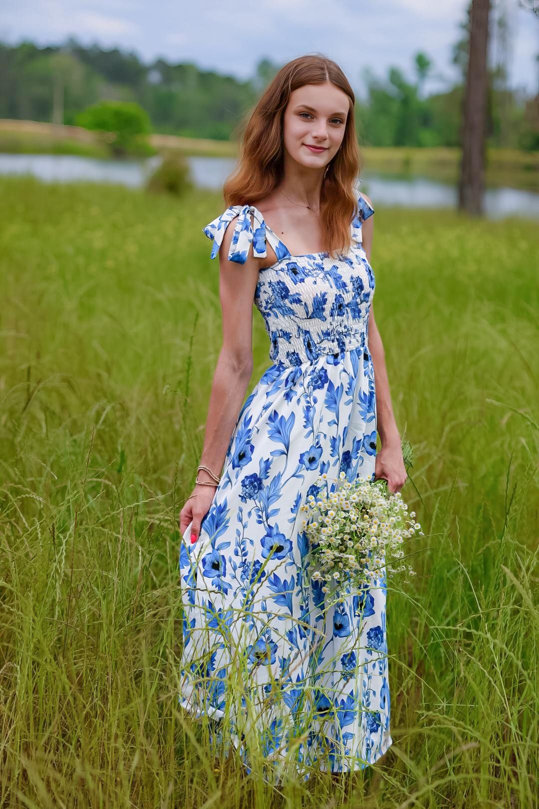 Young woman in blue floral maxi dress holding wildflowers in a green field.