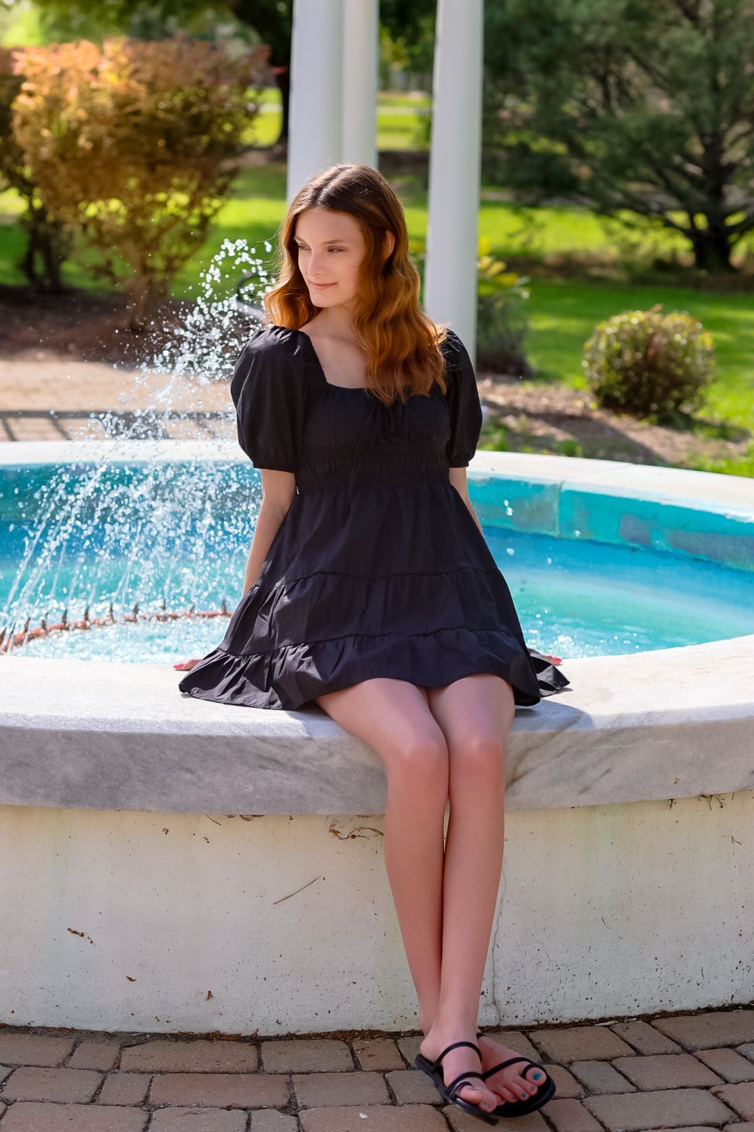 Young woman in black dress sitting on edge of outdoor fountain in lush garden setting.