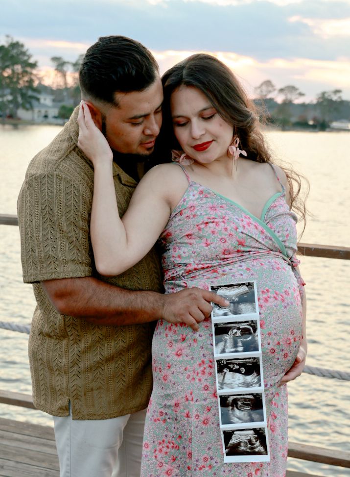A couple shares an intimate moment by the water at sunset while holding ultrasound photos of their expected baby.