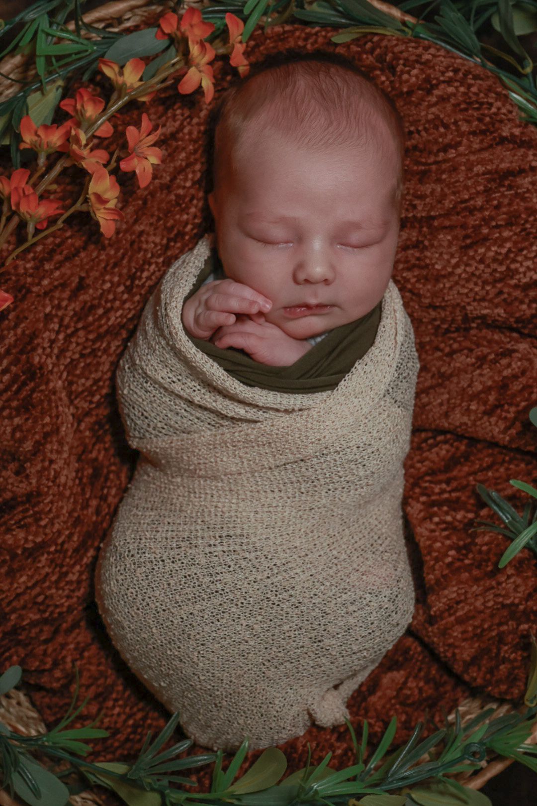 Sleeping newborn baby swaddled in beige wrap on brown textured blanket with orange flowers and greenery as decoration.