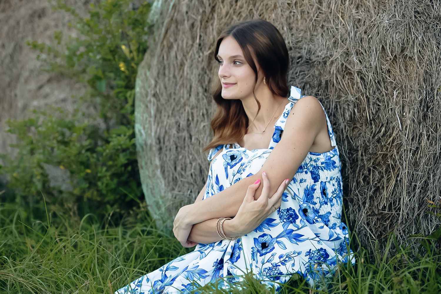 A person in a blue and white floral dress poses against a tree trunk in a natural outdoor setting.
