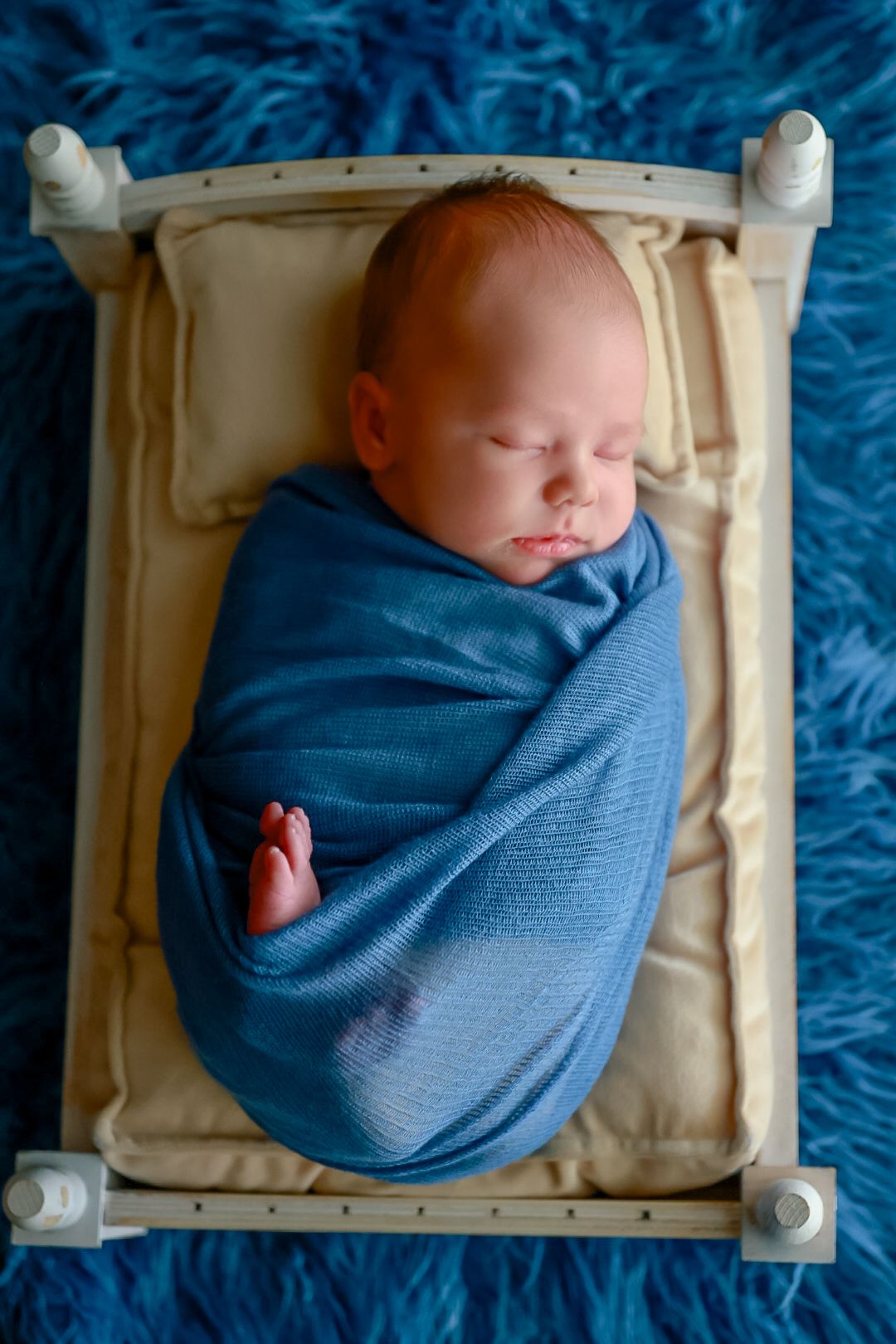 A newborn baby swaddled in a blue blanket rests peacefully in a white wooden crate on a blue textured background.