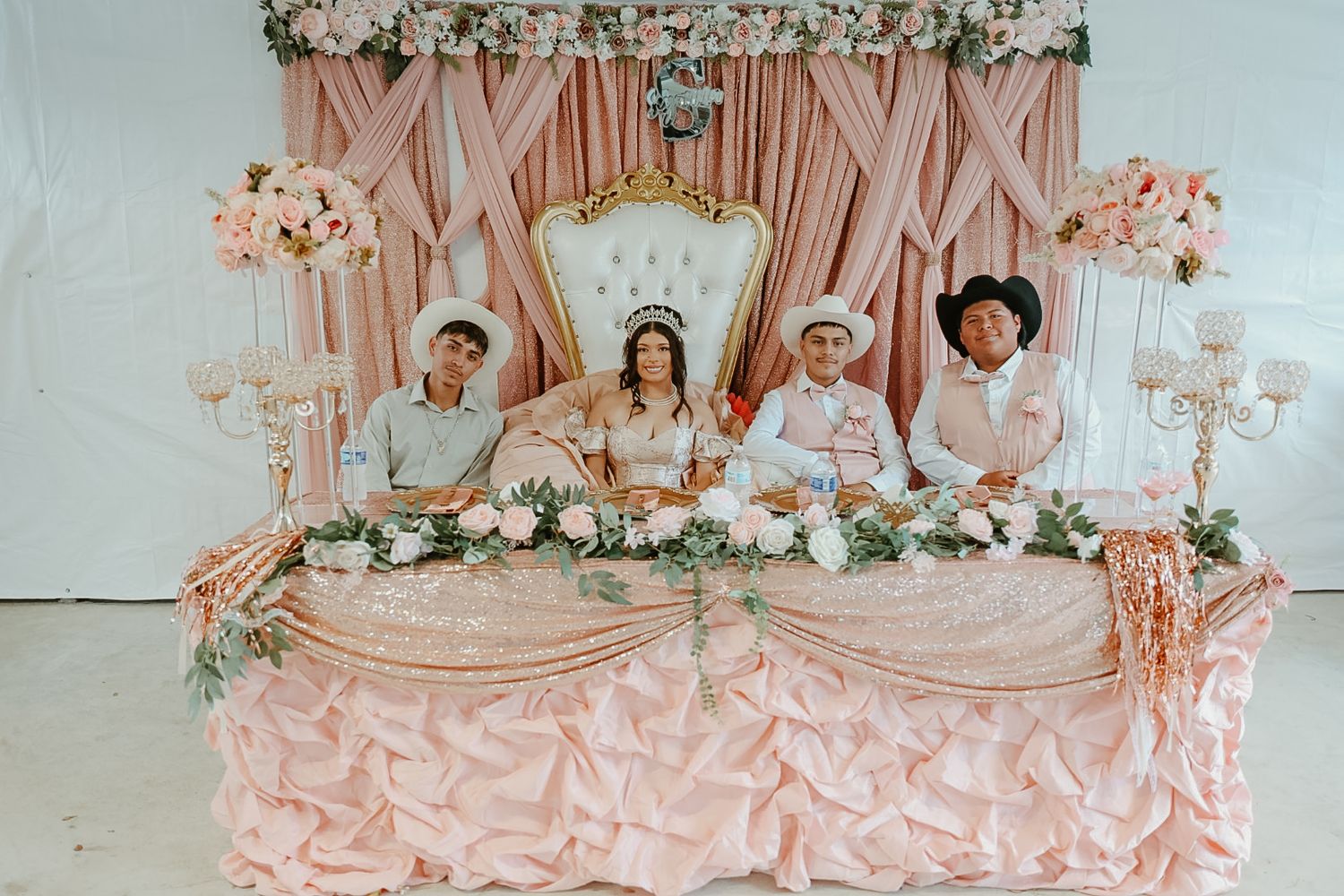 A girl in a tiara sits on a gold throne with three boys at a pink floral quinceañera head table.