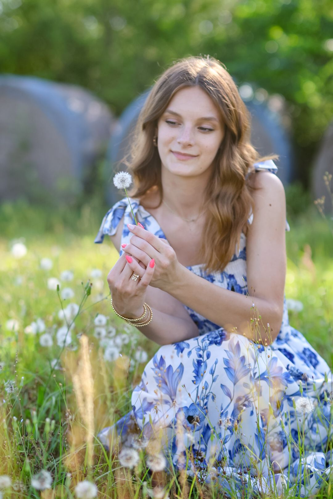 Young woman in blue floral dress holding dandelion in green meadow.