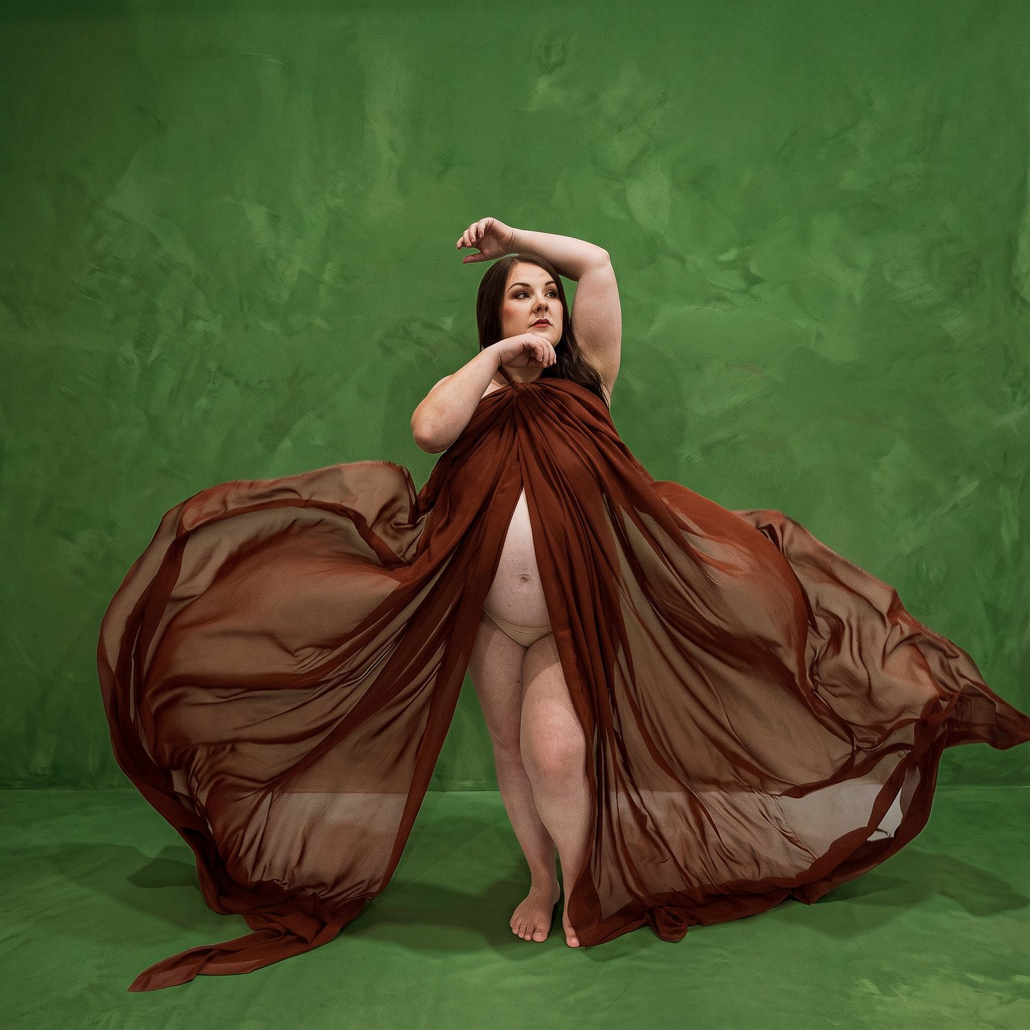 Plus-size woman poses gracefully in flowing rust-colored fabric against a green backdrop.