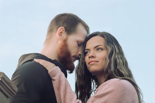A young couple sharing an intimate moment outdoors, the man with a beard leaning close to a brunette woman in a pink top.