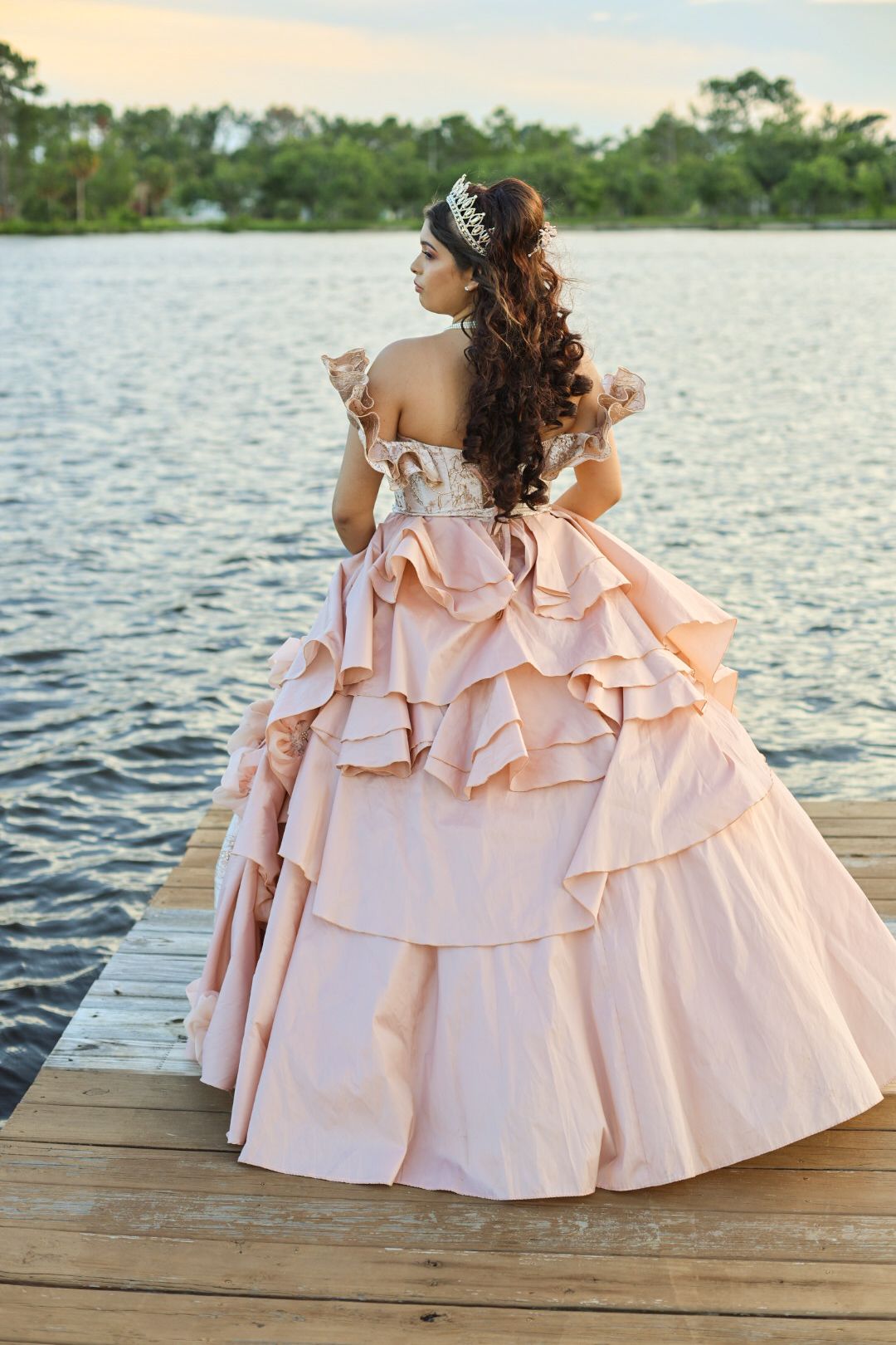 Young woman in blush pink tiered ballgown and crown stands on dock overlooking serene lake at sunset.
