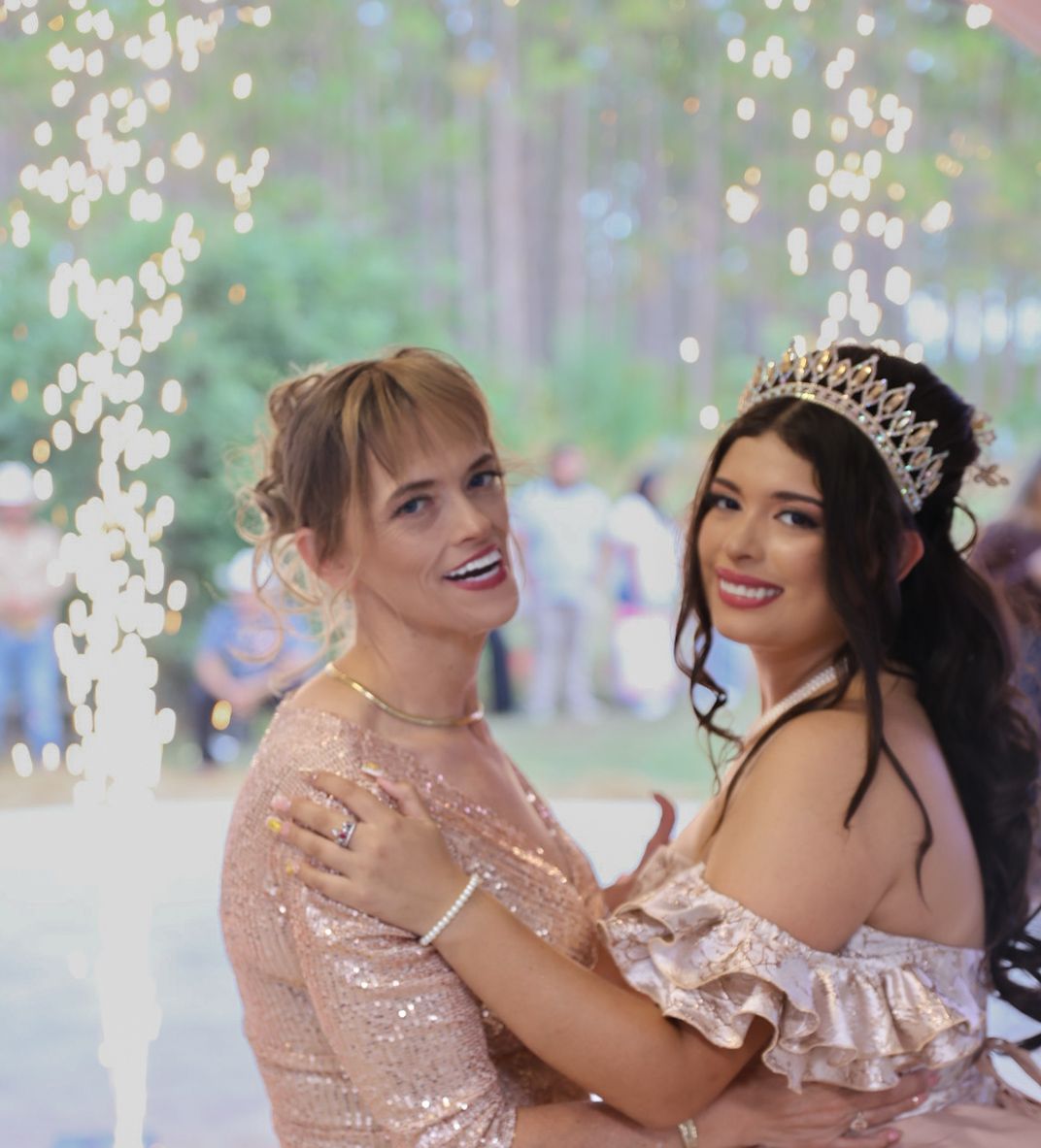 Two young women in elegant dresses pose together at a quinceañera celebration, one wearing a sparkling tiara.