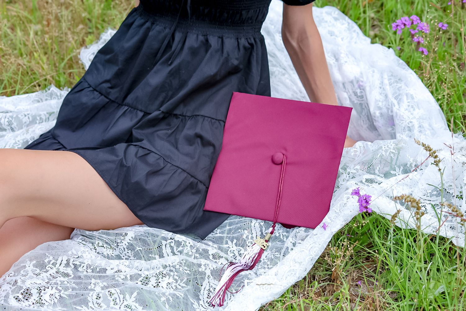 Graduation cap and gown laying on grass with pink flowers in the background.