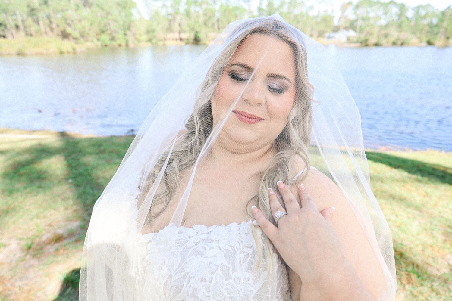 Bride in white veil poses outdoors by a lake on a sunny day with natural scenery.