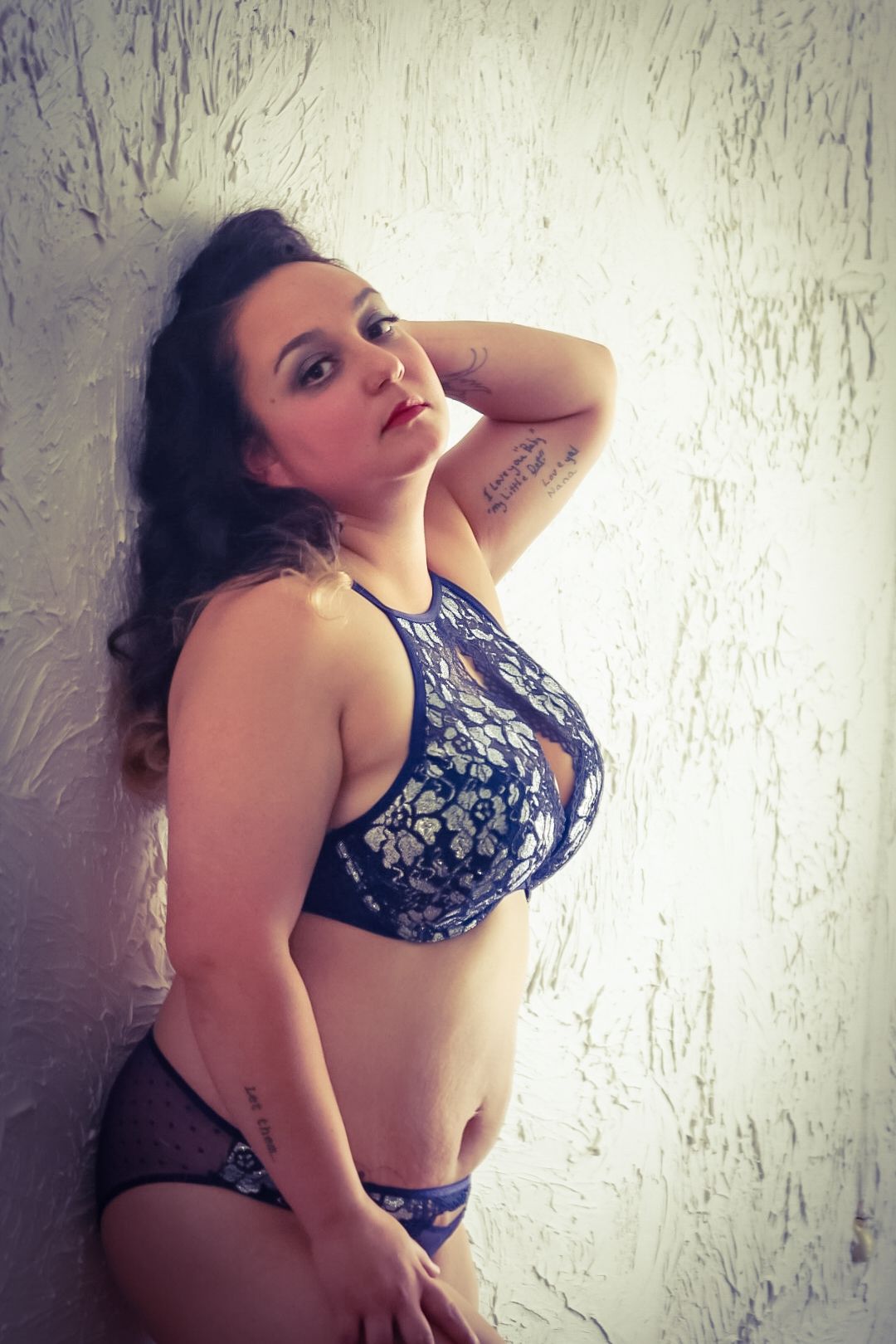 Young woman in blue lace lingerie posing against a textured white wall with tattoos visible on her arm.