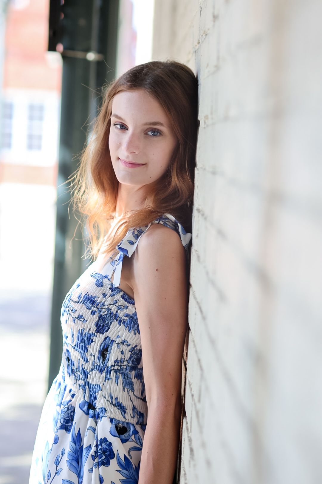 Young woman in blue floral sundress leaning against white brick wall outdoors.