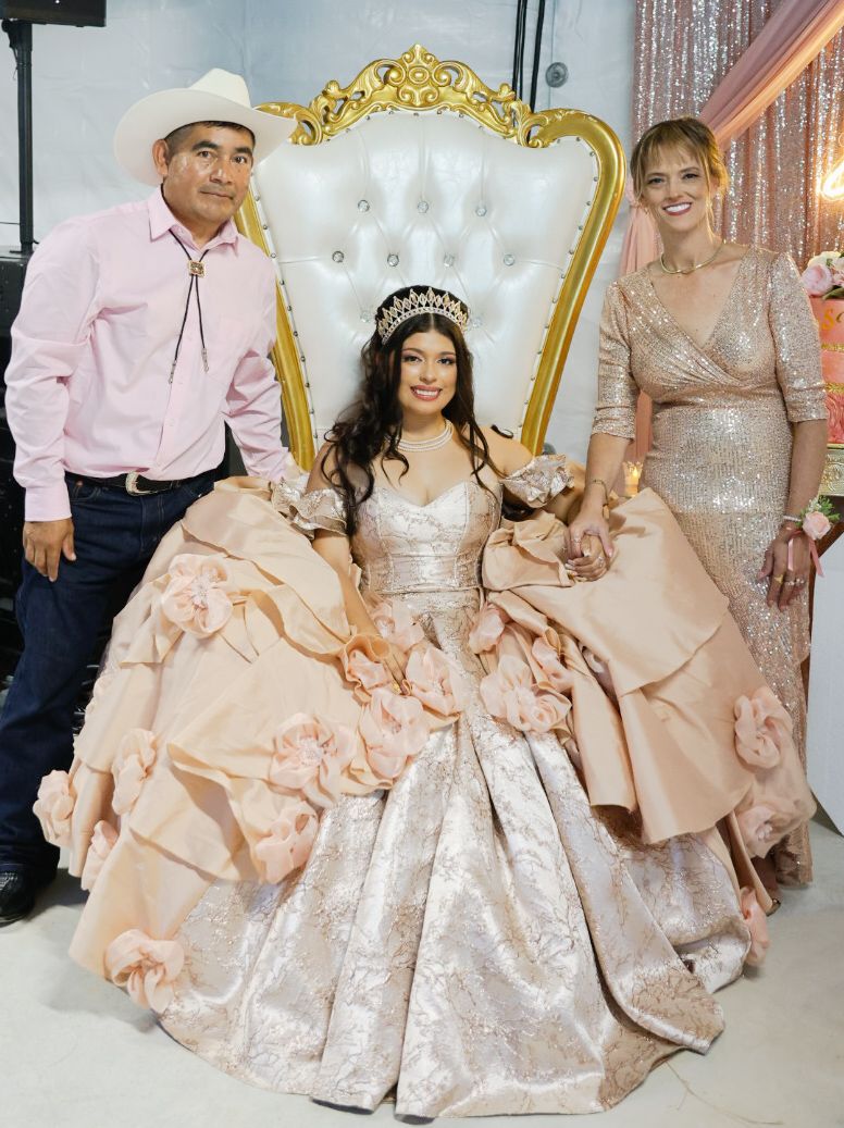 Young woman in pink quinceañera gown seated on ornate throne chair flanked by proud parents at celebration.