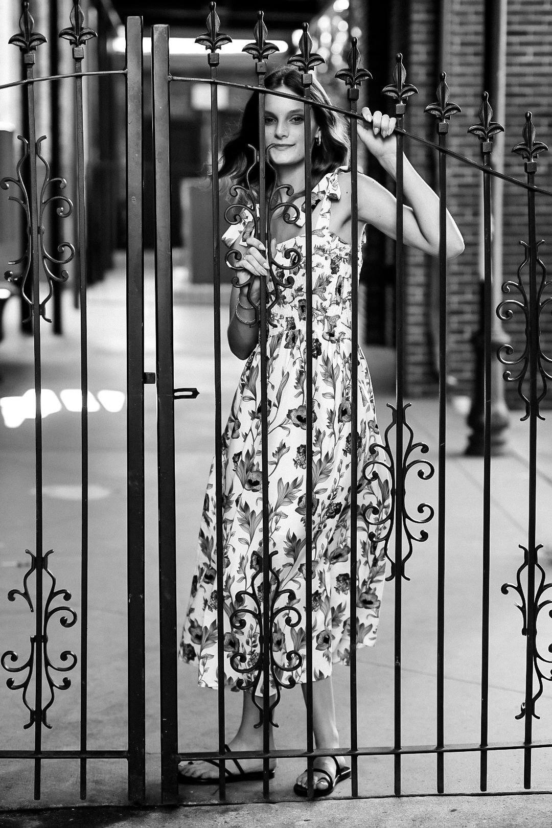 Black and white photo of a woman in a floral dress standing behind ornate iron gates.