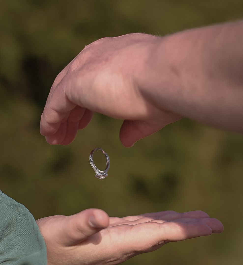 A diamond engagement ring being dropped into an open palm against a blurred green background.