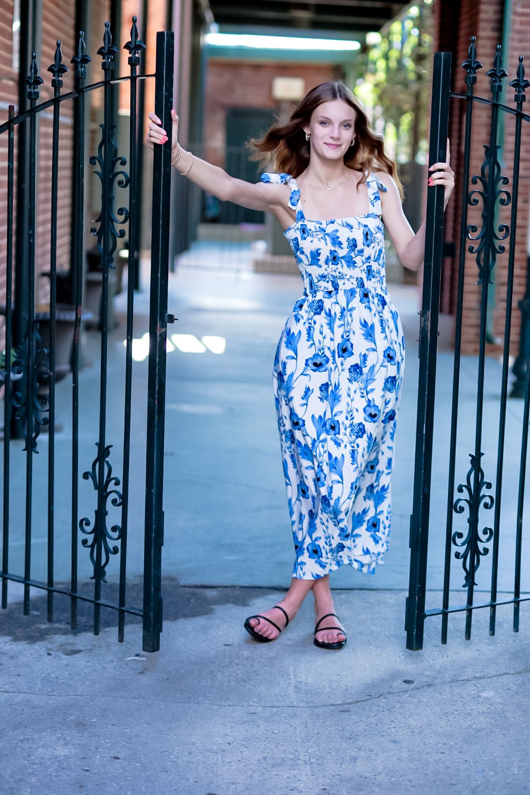 Woman in blue floral midi dress standing between black iron gates on a city sidewalk.