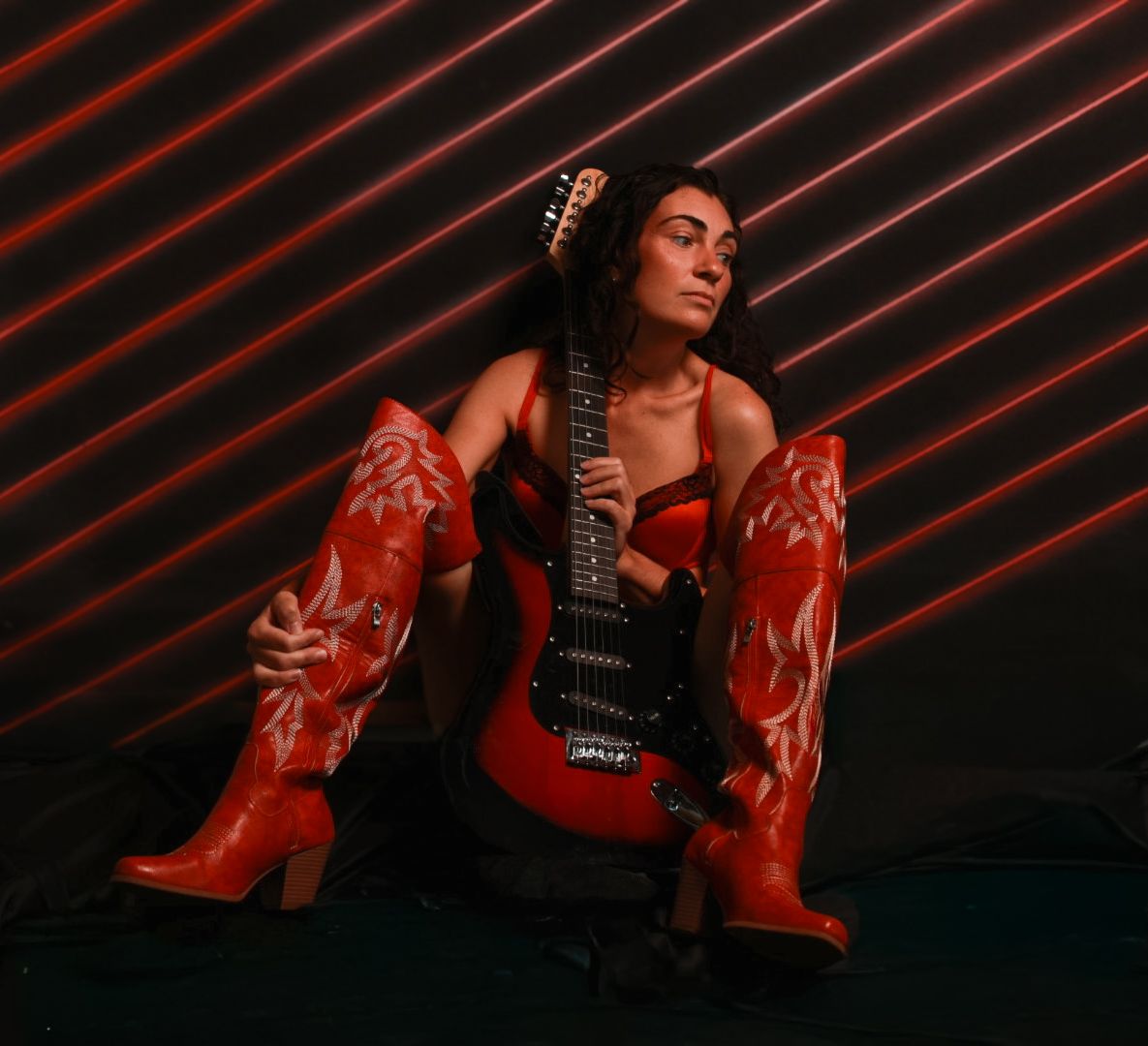 Woman in red cowboy boots and corset poses with a red electric guitar against a neon-lit striped background.