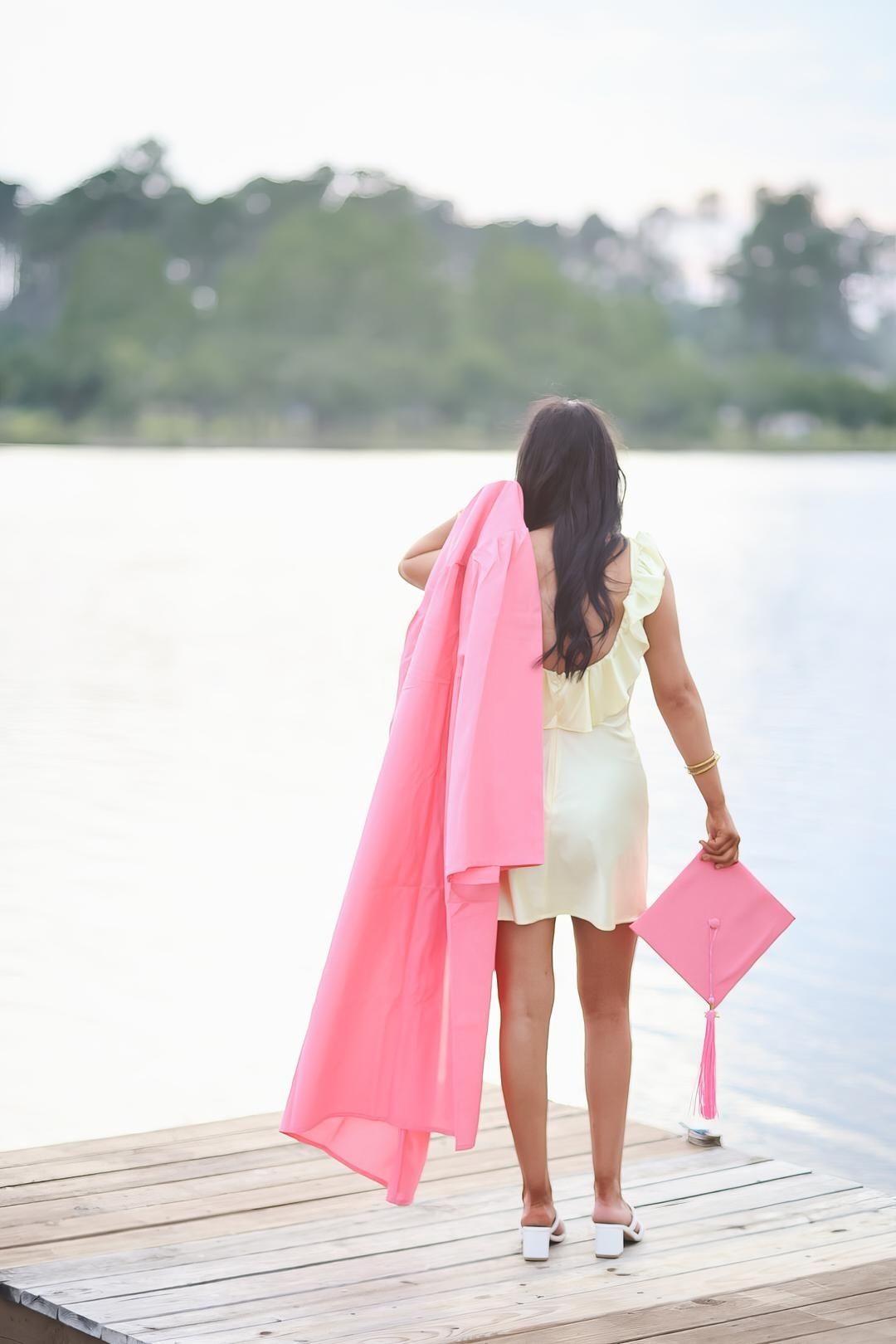 Graduate in white dress and pink graduation gown stands on wooden dock overlooking water.