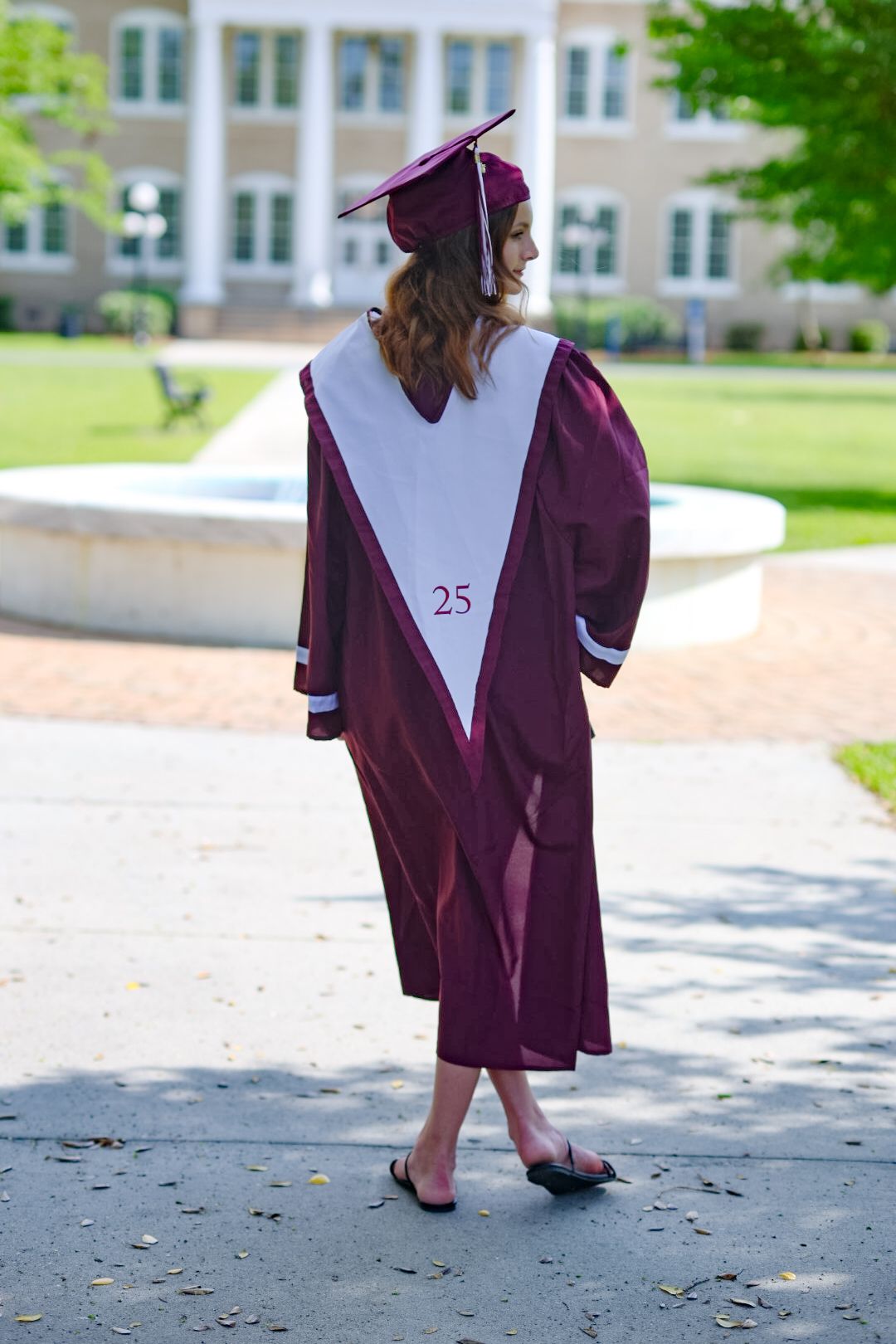 Female graduate in maroon cap and gown viewed from behind on campus walkway.