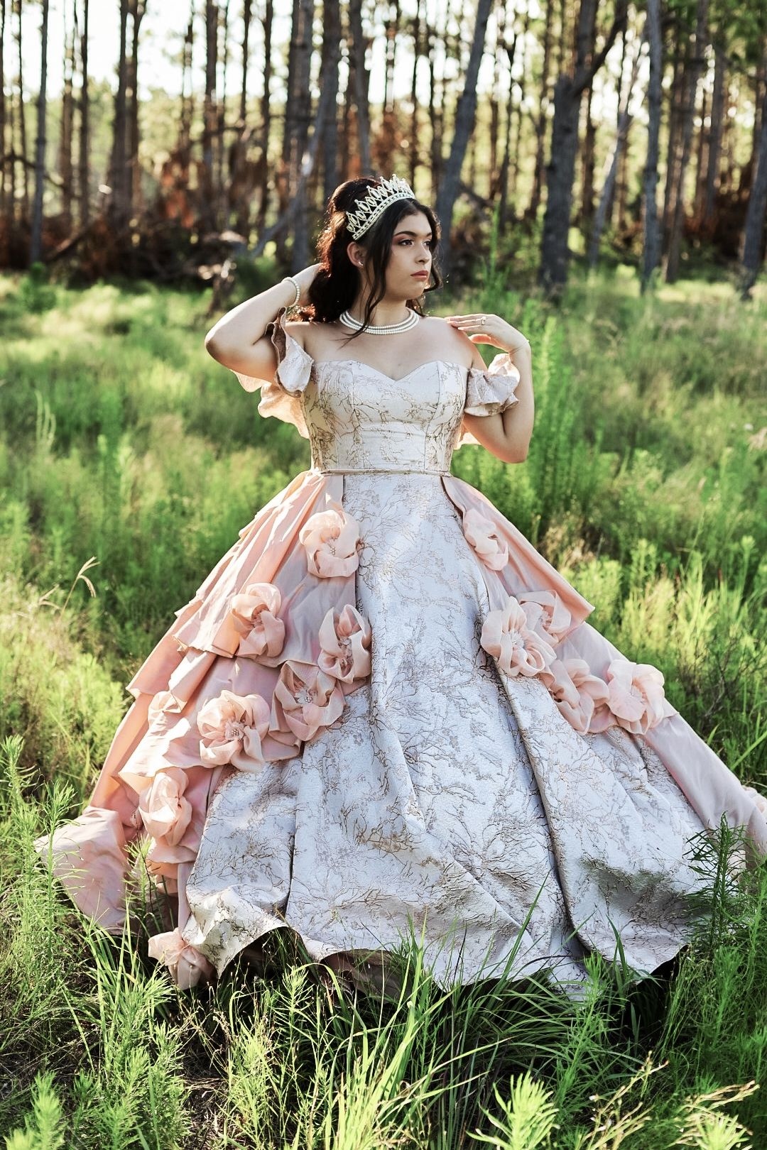 Woman in ornate pink and white ball gown with floral details and tiara poses gracefully in a sunlit forest.