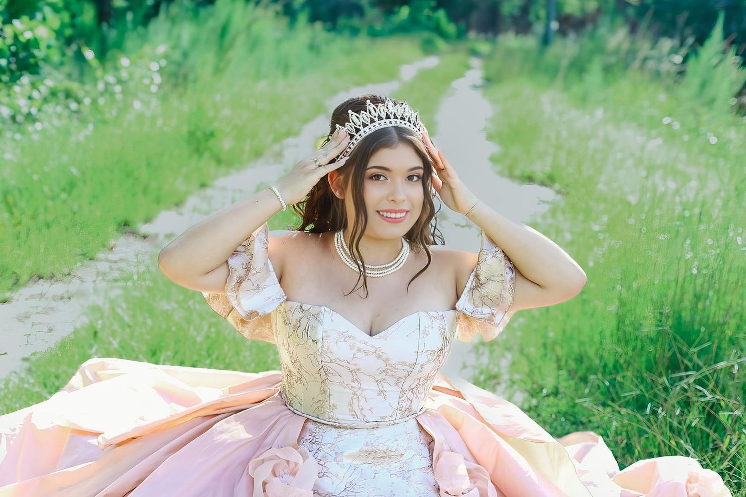 Young woman in pink ball gown and tiara poses on a garden path surrounded by lush greenery.