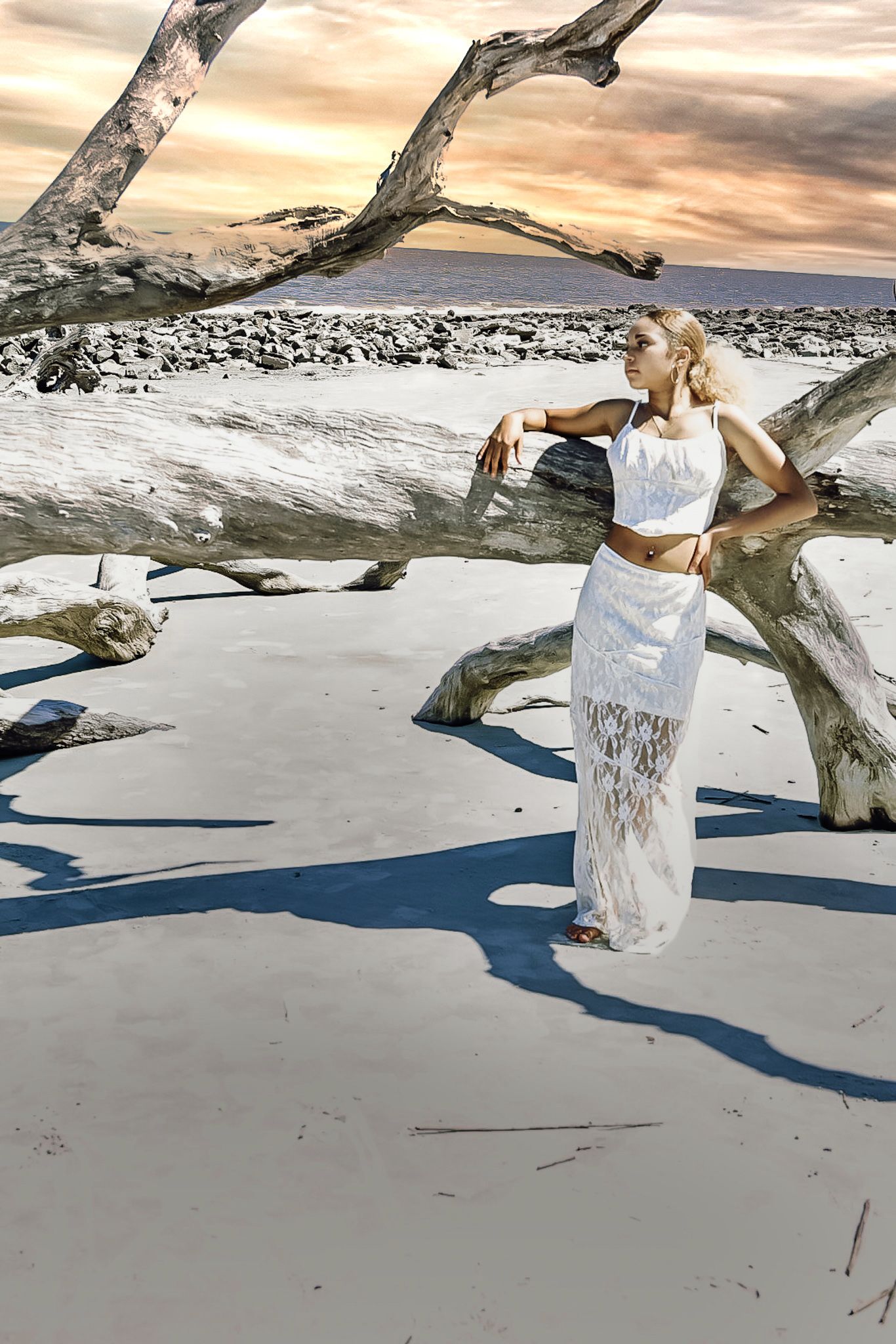 A figure in a white lace outfit poses on a driftwood-strewn beach at sunset with dramatic cloudy skies.