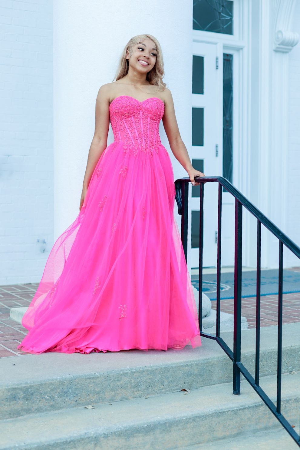 A person in a vibrant hot pink strapless ball gown poses elegantly on white stone steps with a black railing.
