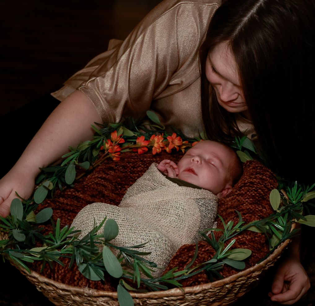 Newborn baby wrapped in cream knit blanket rests in brown basket adorned with greenery and orange flowers.