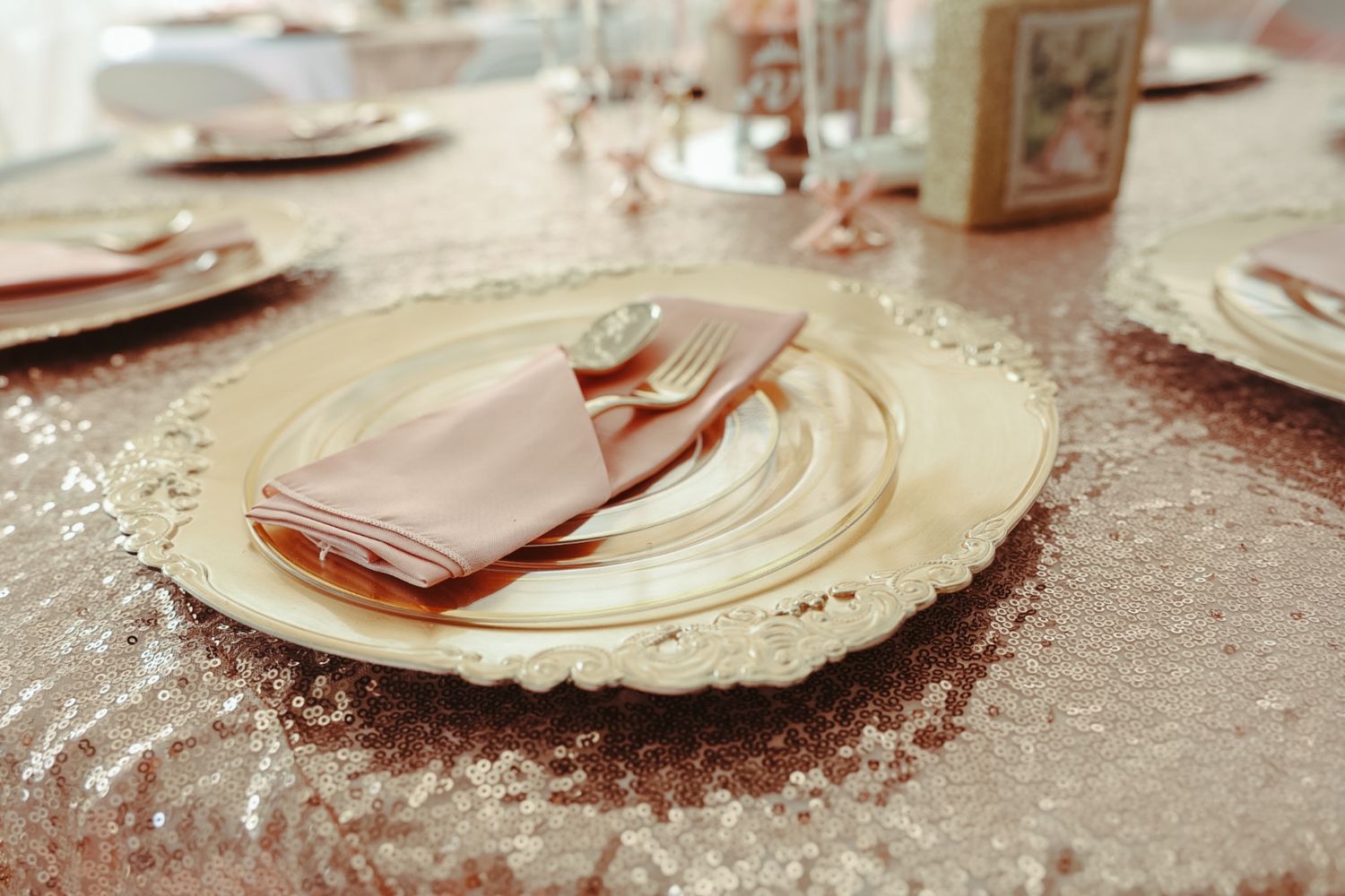 Elegant rose gold sequin tablecloth with ornate charger plate, pink napkin, and gold cutlery for a wedding reception.