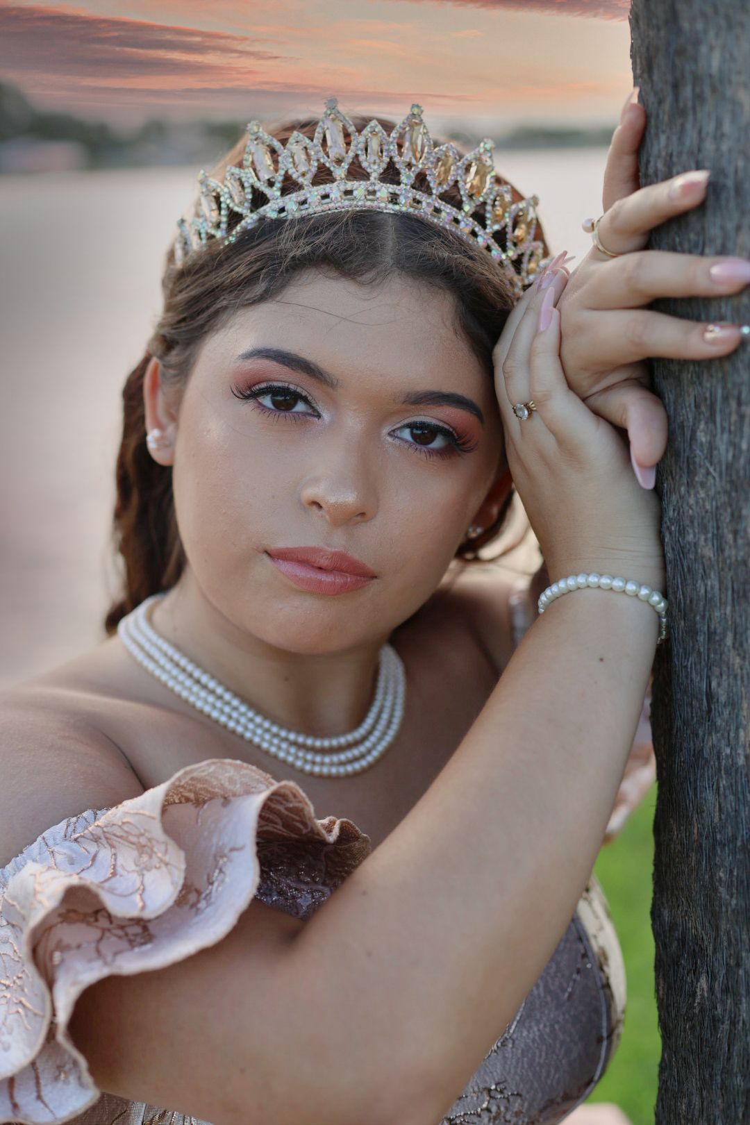 Young woman in tiara, pearl necklace, and bracelet leans against tree at sunset in elegant gown.
