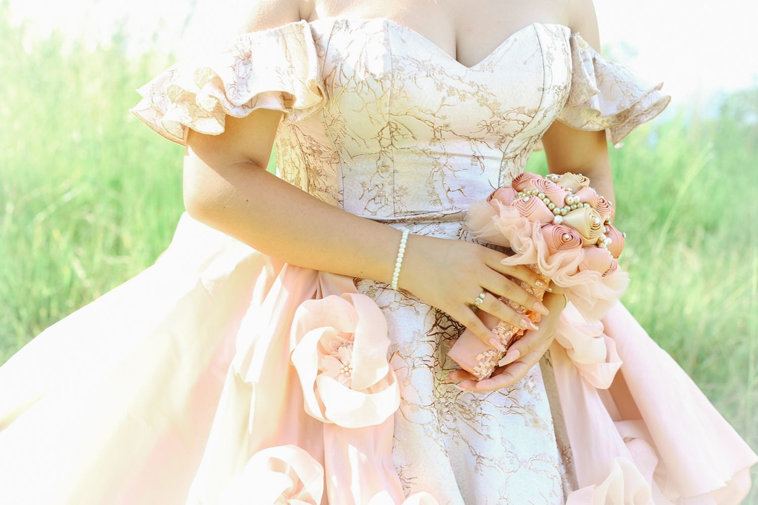 Woman in elegant off-shoulder pink ball gown holding a blush floral bouquet in a lush green field.