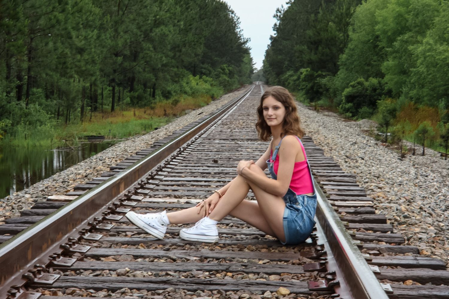 Young woman in denim shorts sitting on railroad tracks surrounded by lush green forest trees.