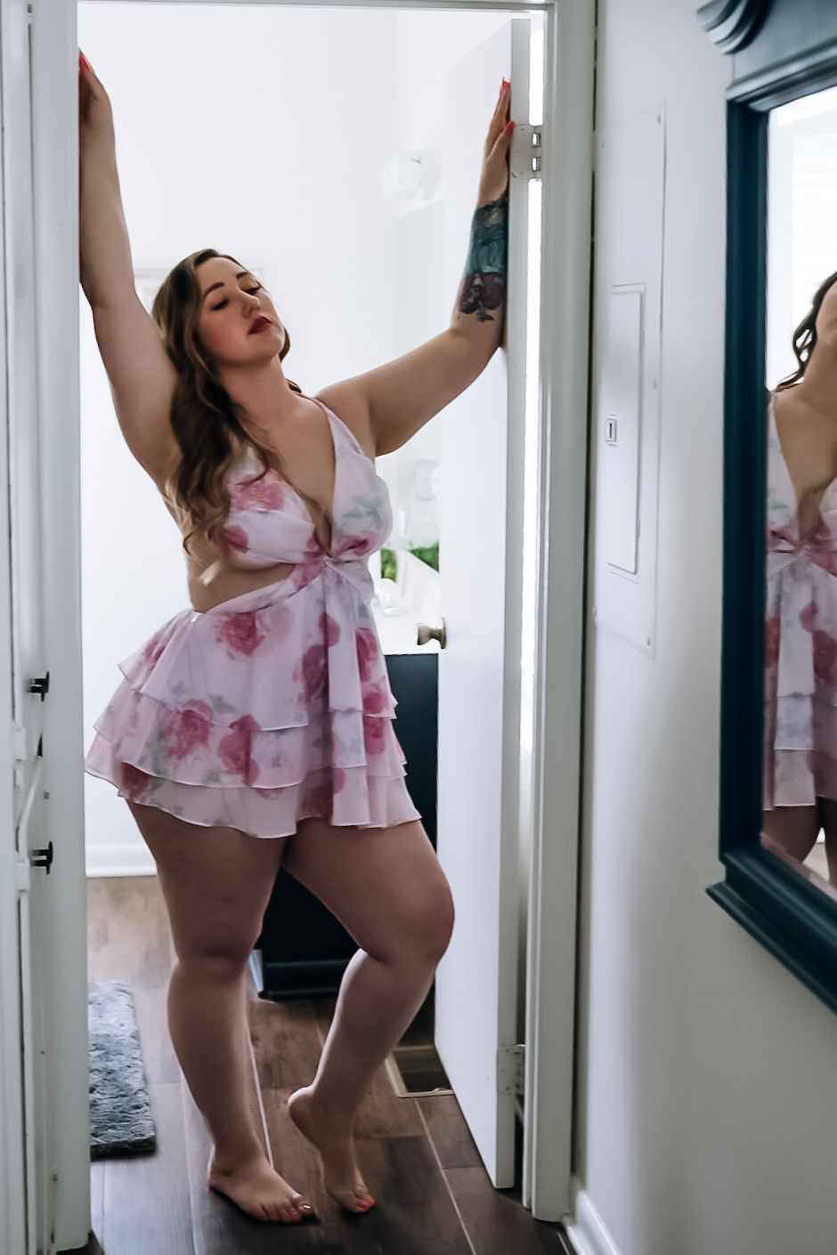 Woman in floral pink babydoll lingerie posing in doorway with arms raised, mirror reflection visible.