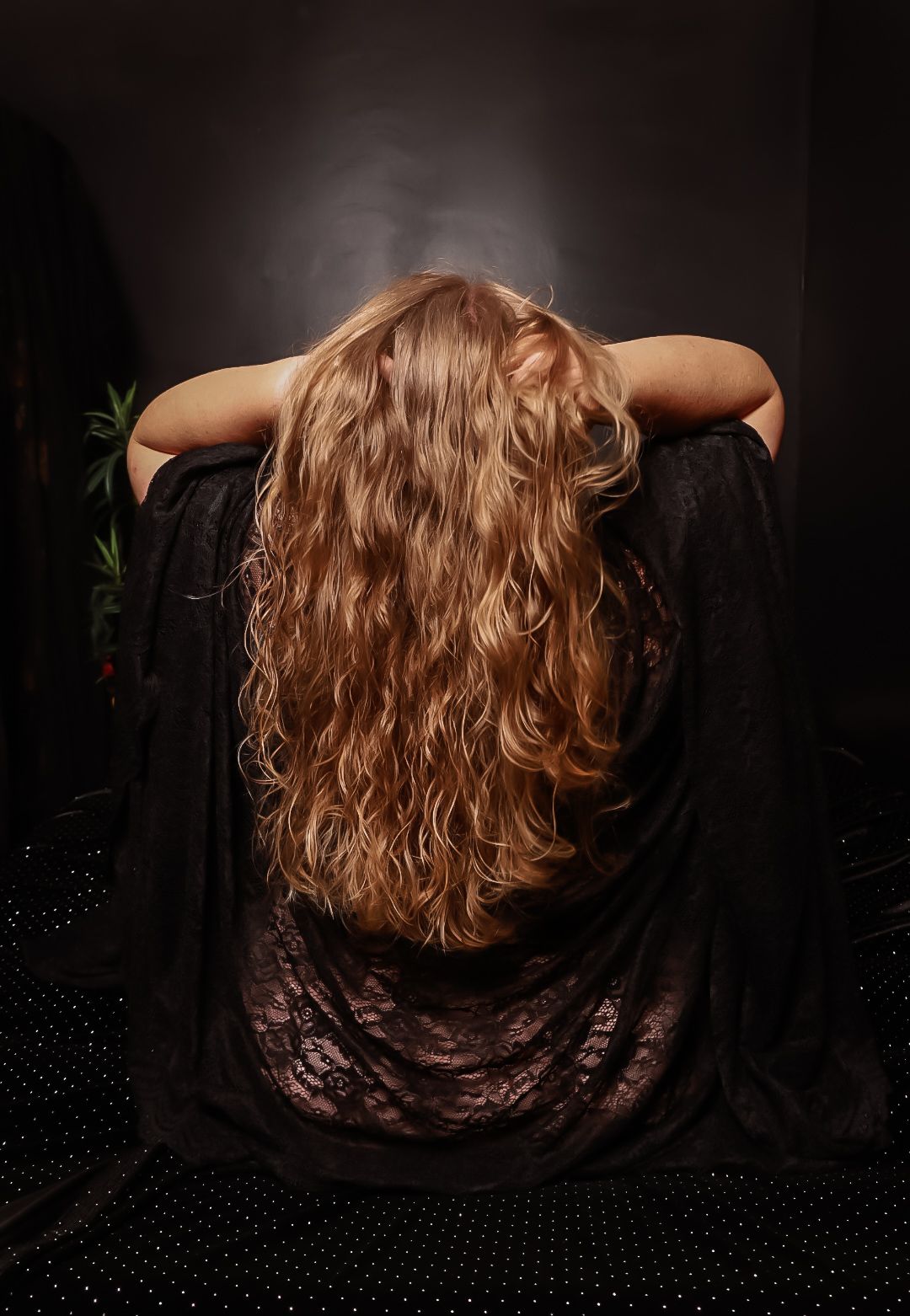 Woman with long, wavy auburn hair wearing black lace, photographed from behind against a dark background.