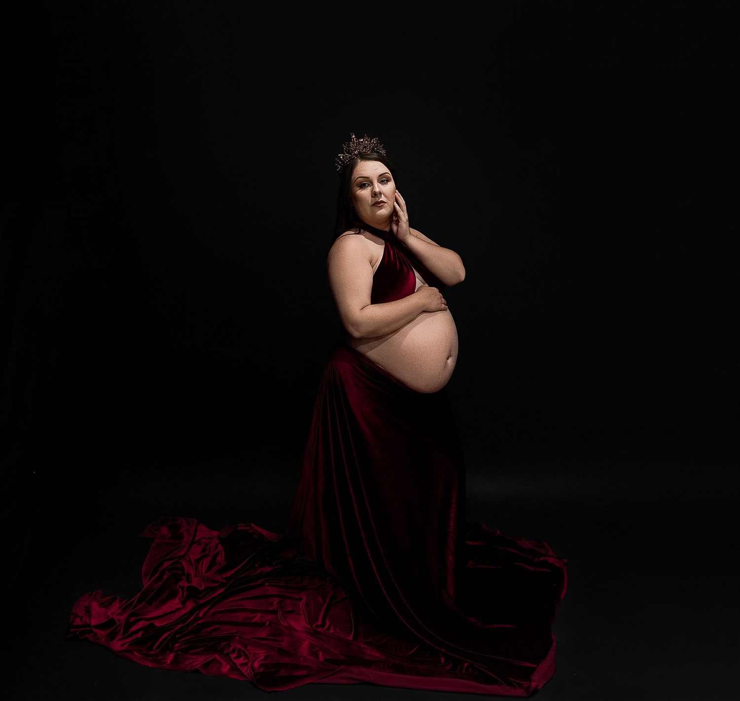 Pregnant woman in red velvet gown and crown poses gracefully against dark background in elegant maternity portrait.