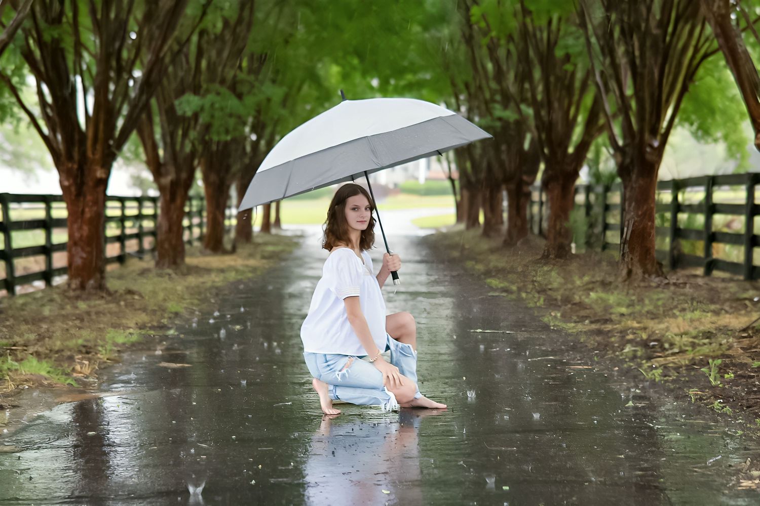 Woman and child sitting on wet road under umbrella surrounded by tree-lined path in the rain.